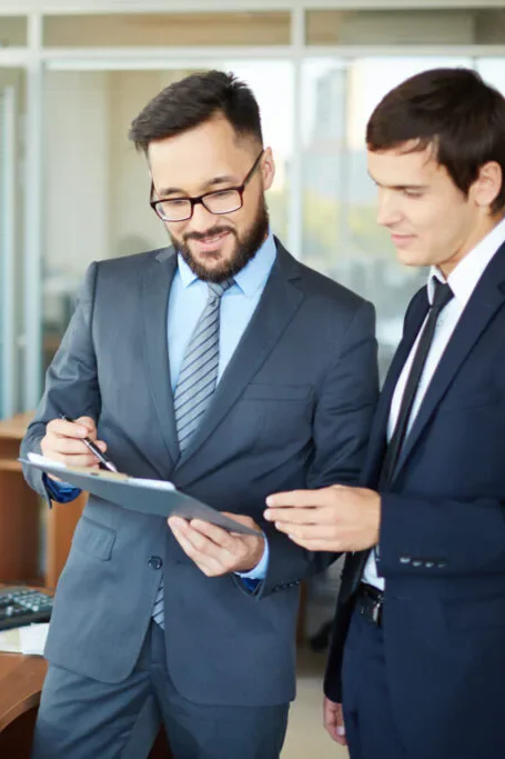 Two businessmen in suits discussing documents in an office setting.