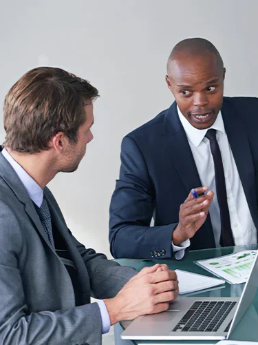 Two men in business suits having a serious discussion in an office, with laptops and documents on the table.