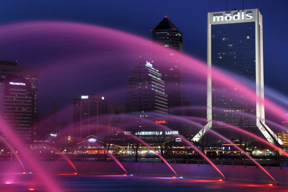 Nighttime view of the Jacksonville, FL, skyline with illuminated buildings and a fountain with pink and purple lights in the foreground.