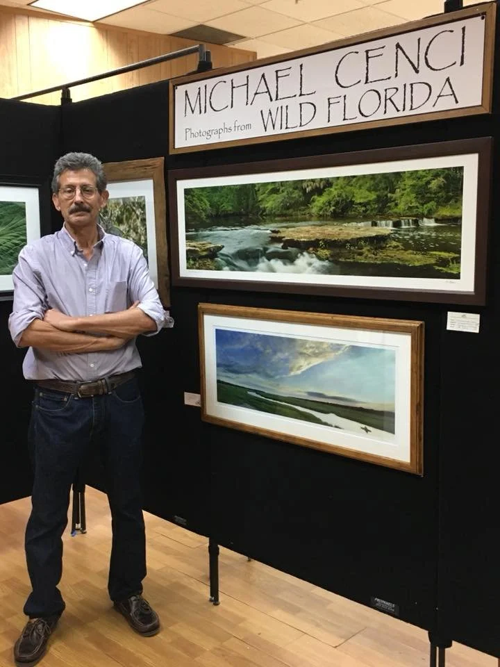 A man with glasses and a mustache, wearing a light purple shirt with rolled-up sleeves, black pants, and brown shoes, stands with crossed arms next to a display of landscape photographs at an art exhibit titled 'Michael Cenci Wild Florida.' The display features three framed photographs of natural outdoor scenes, including a river scene with rocks and greenery, and a landscape with a cloudy sky and water.