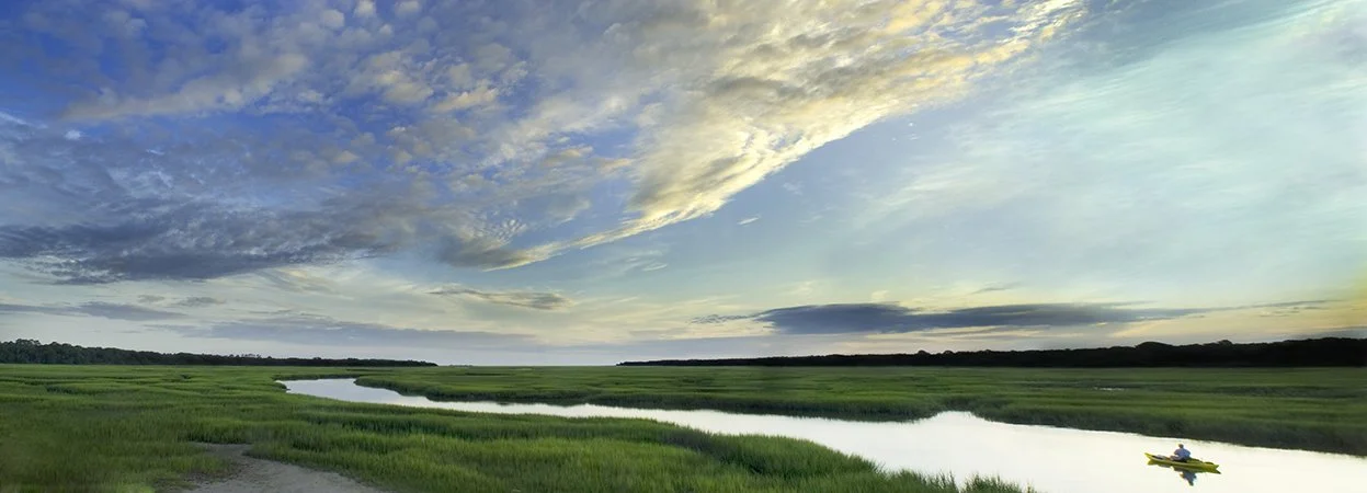 A person kayaking on a river flowing through green marshland under a partly cloudy sky.