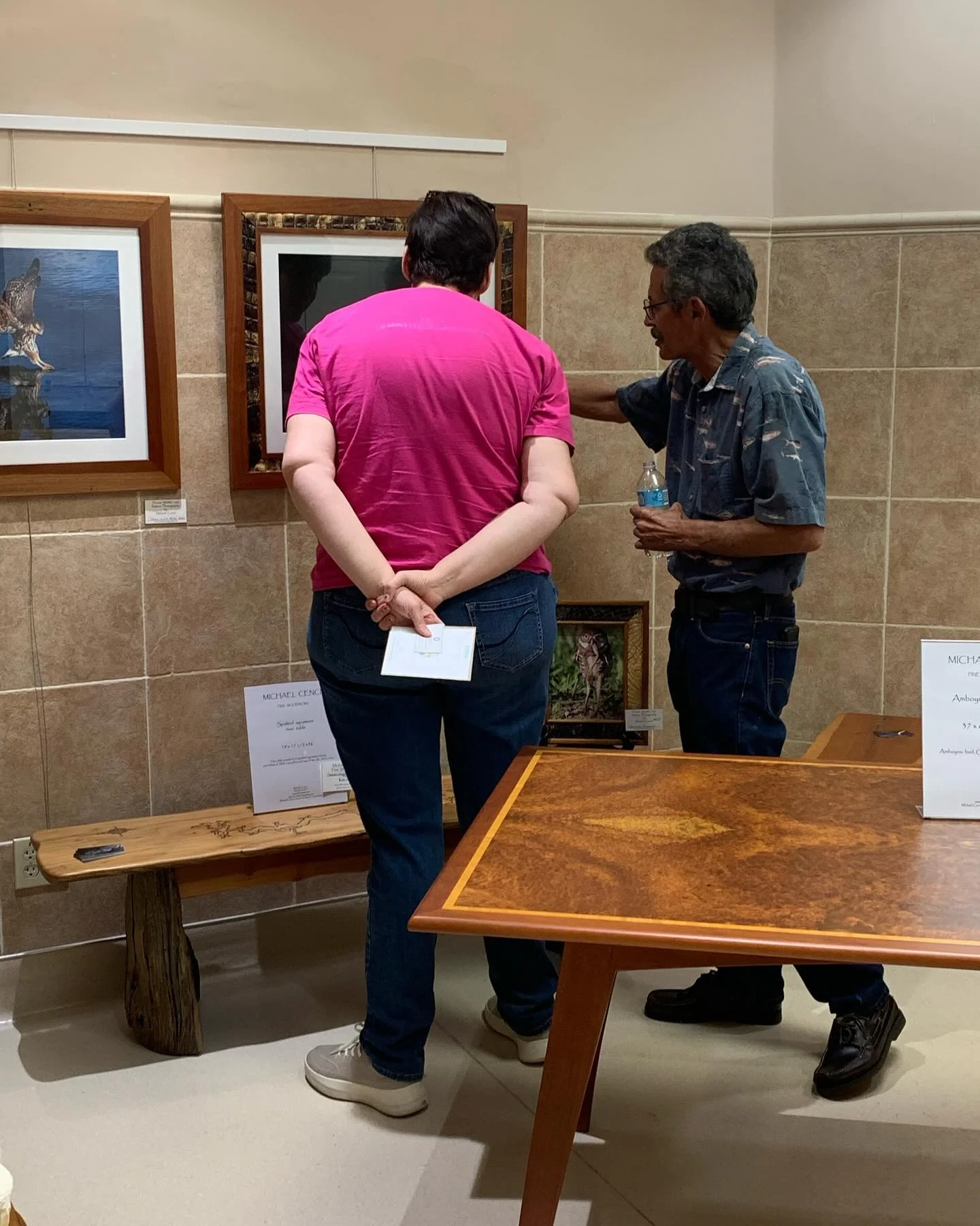 Two people viewing artwork in an art gallery, one person in a pink shirt holding a pamphlet and another in a blue shirt holding a water bottle, engaging in conversation.