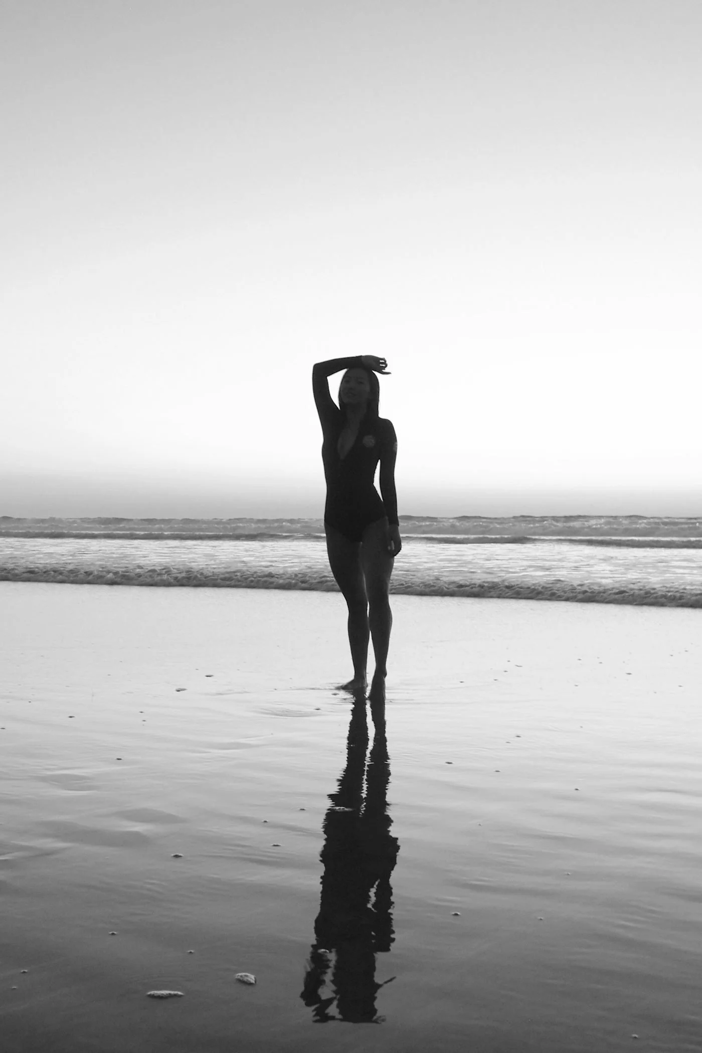 Silhouette of a woman in a swimsuit standing on the beach with her reflection in the wet sand, ocean waves in the background, during sunset or sunrise, in black and white.