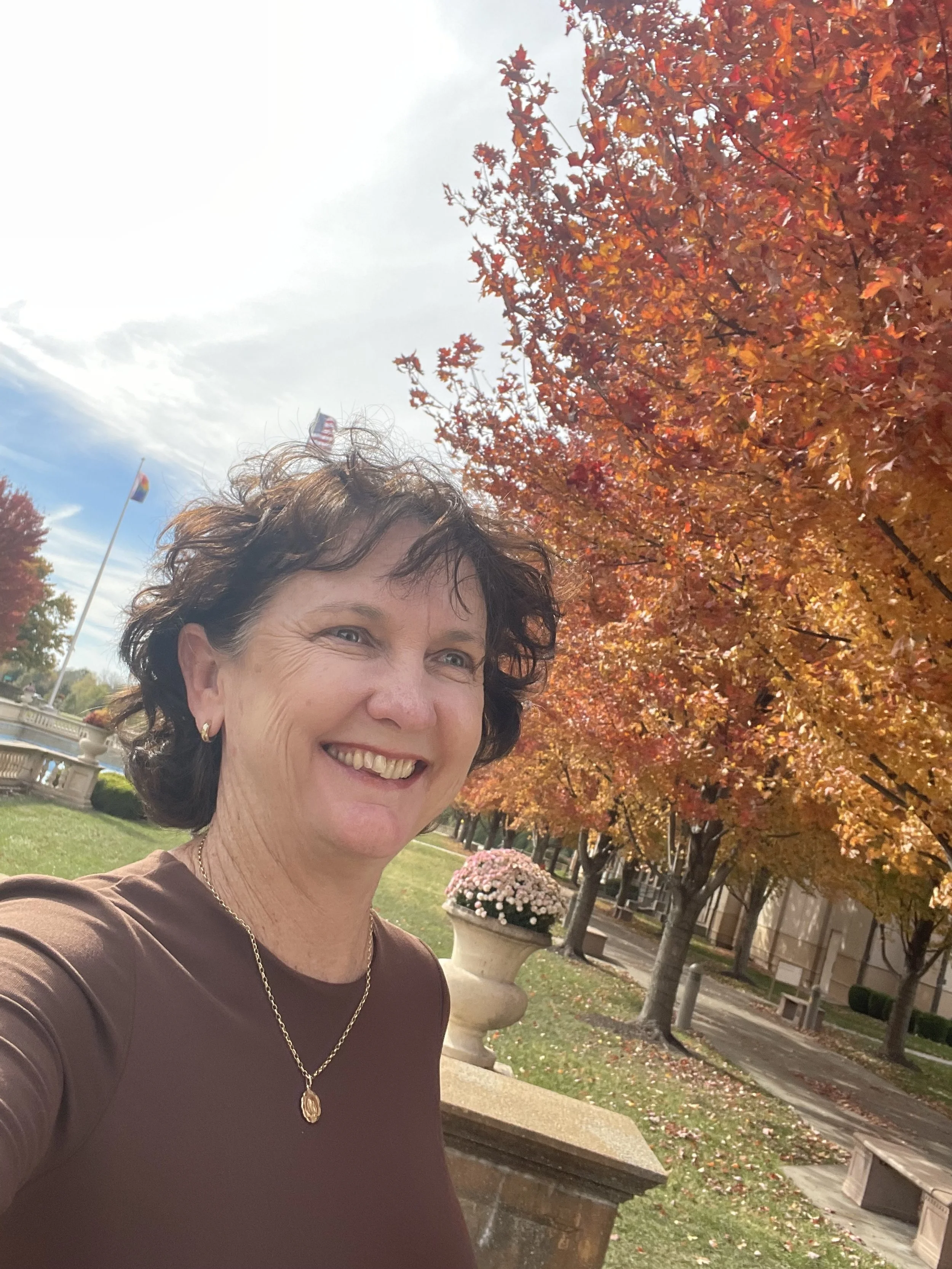 A woman smiling outdoors in front of red and orange autumn trees, with flags and a fountain in the background.
