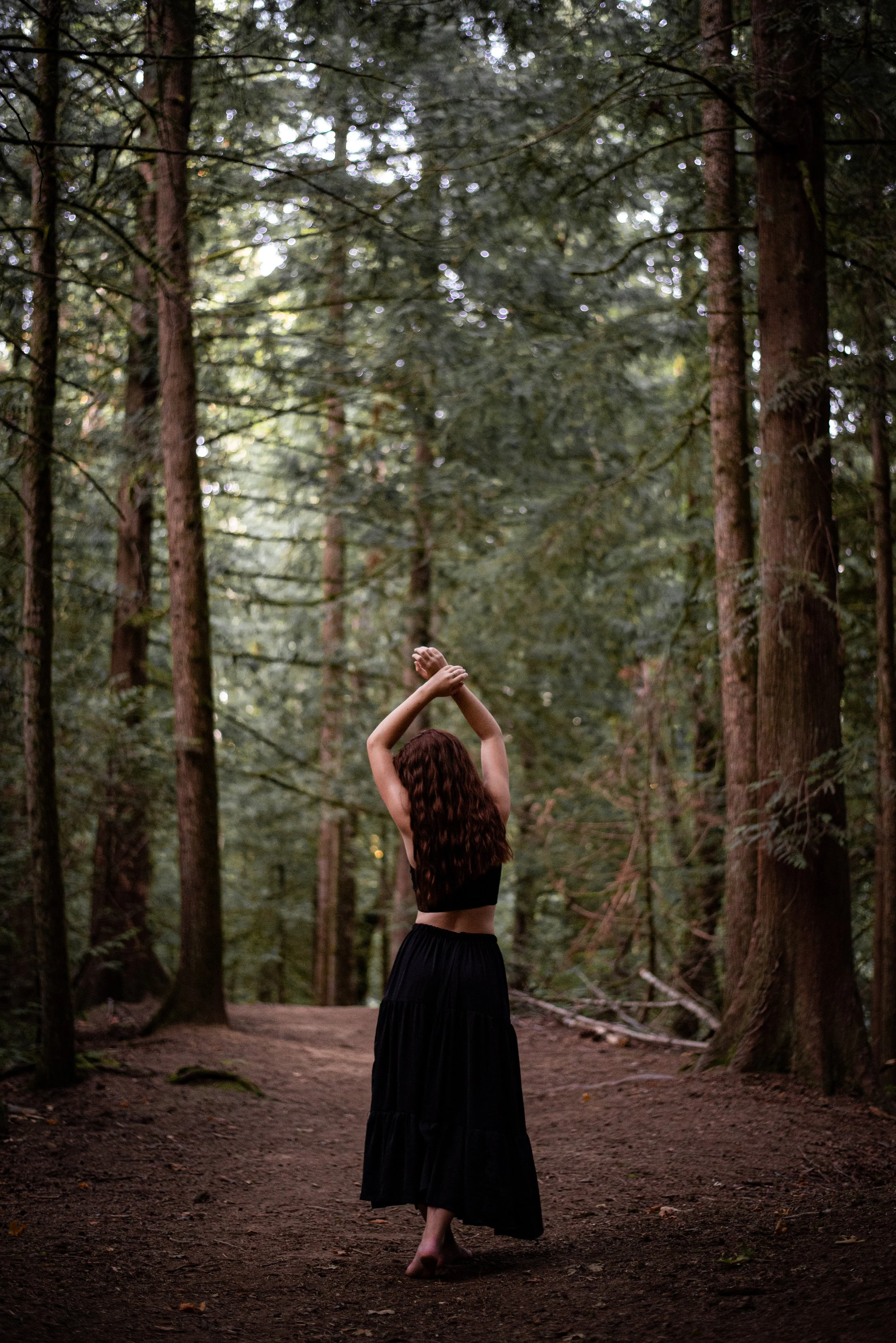 A woman with long, wavy brown hair wearing a black crop top and long black skirt walking barefoot on a dirt trail in a dense forest with tall trees.