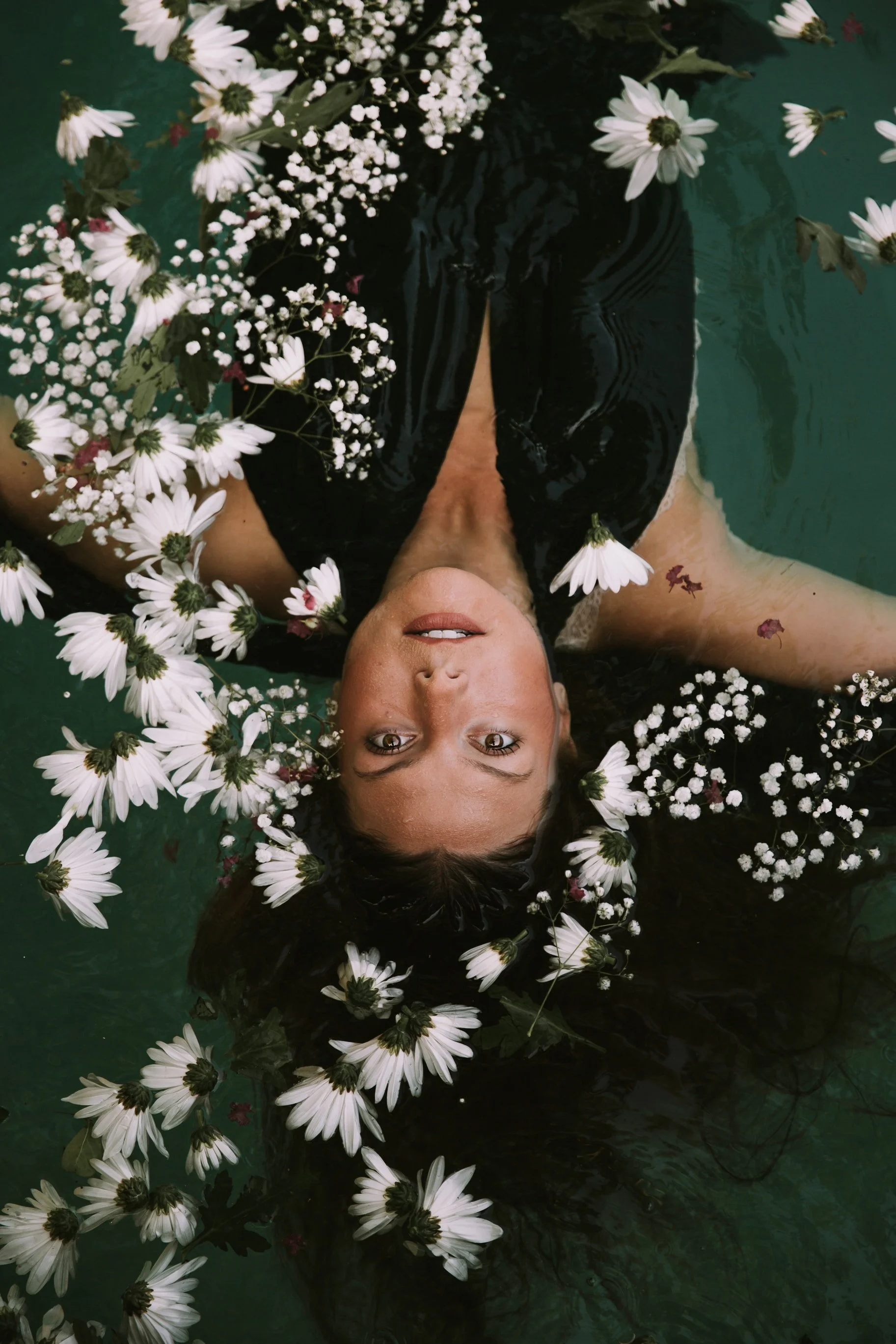 A woman floating on her back in water, surrounded by white daisies and small white flowers, with her head tilted back and eyes looking up.