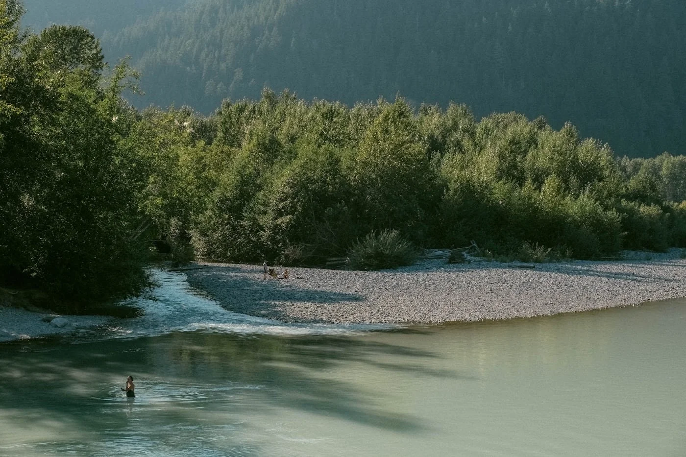 A river flowing through a forested landscape with a person wading in the water and others on the bank, surrounded by trees and distant mountains.