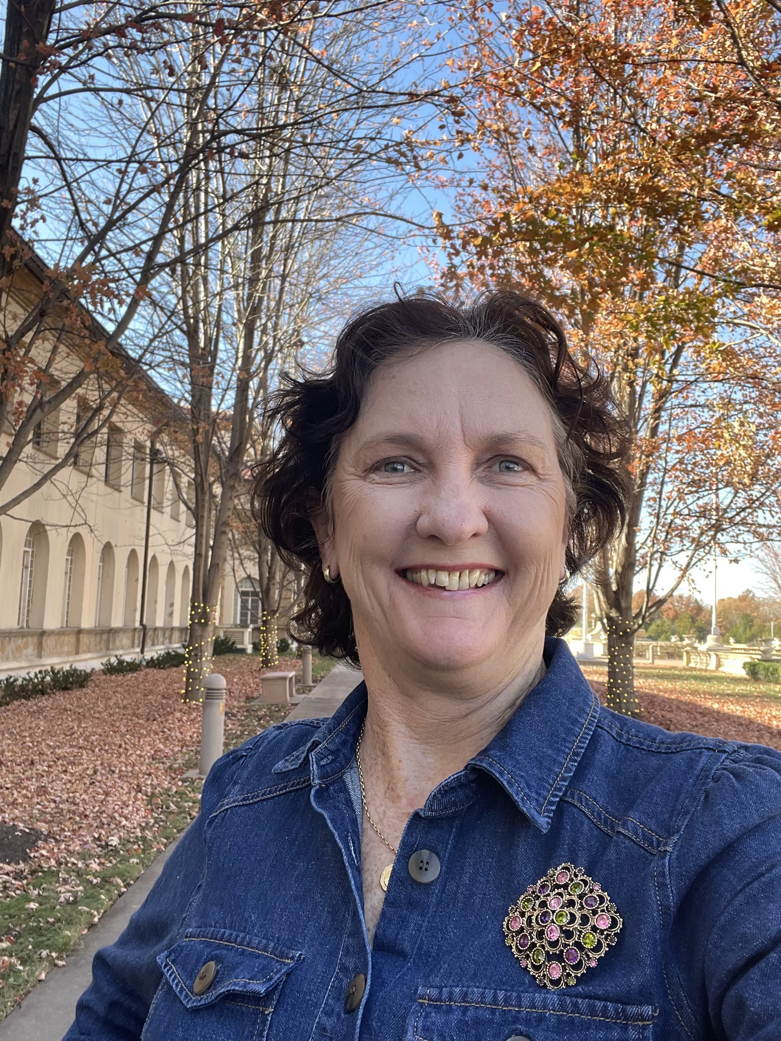A woman taking a selfie outdoors during fall, smiling, with trees and a building in the background. She is wearing a denim jacket and a colorful brooch.