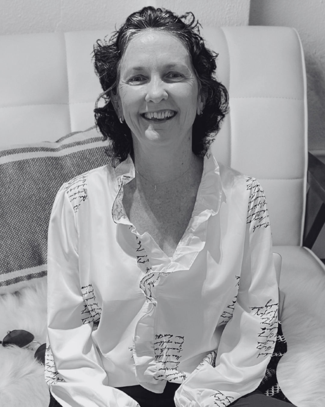 image of Rev. Rachel. A woman sitting on a bed or sofa, smiling at the camera. She has dark, curly hair and is wearing a white blouse with black script writing. There is a pillow and a furry blanket visible behind her.