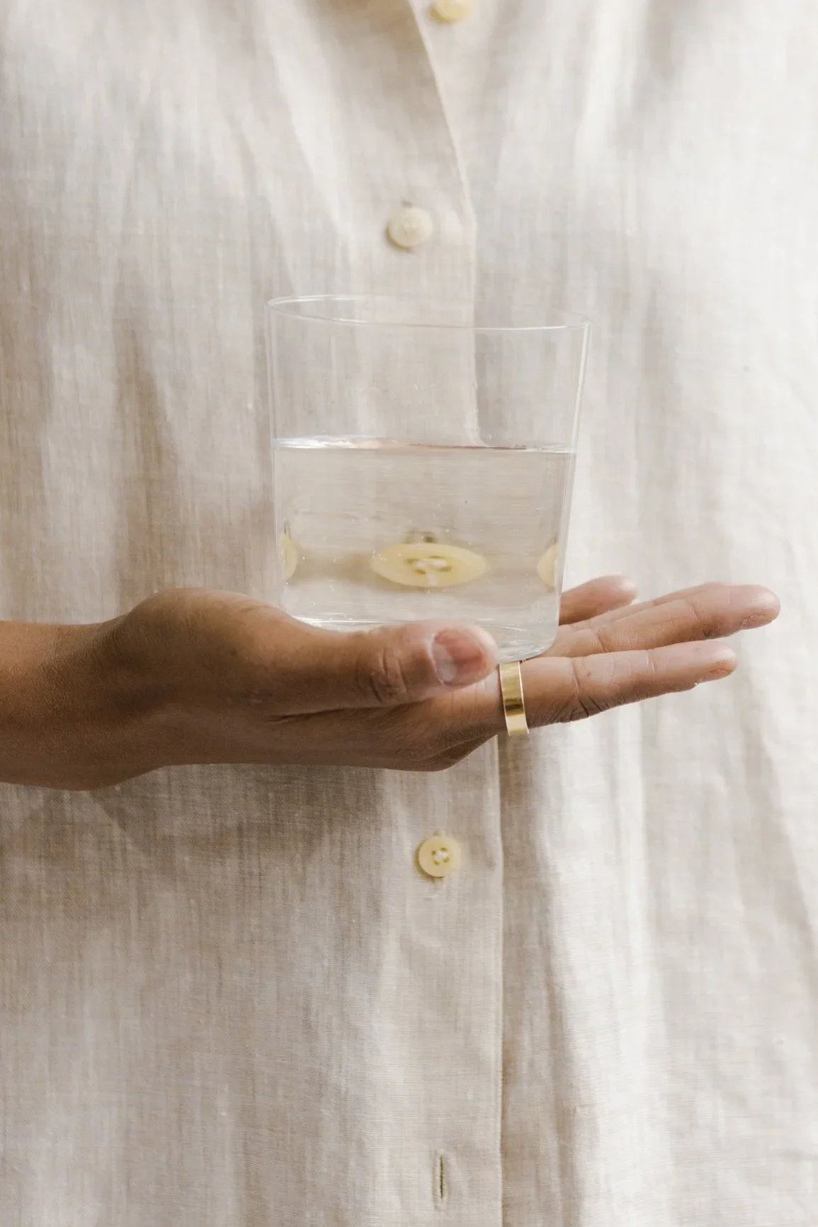 Person wearing a light-colored shirt holding a clear glass of water with slices of banana inside.