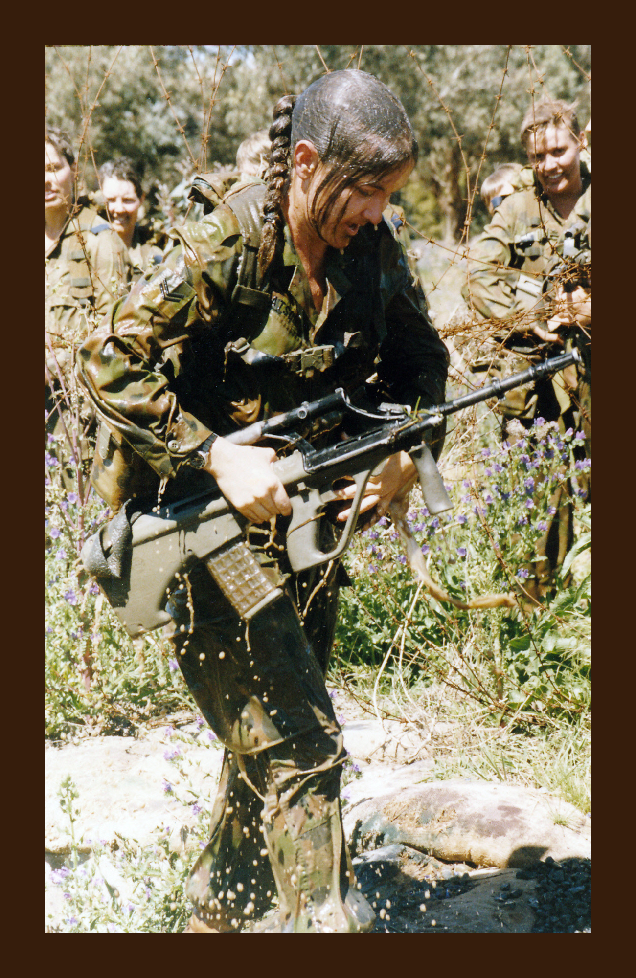 A female soldier in camouflage uniform holding a rifle, surrounded by other soldiers in similar attire in a field with tall grass and flowers.