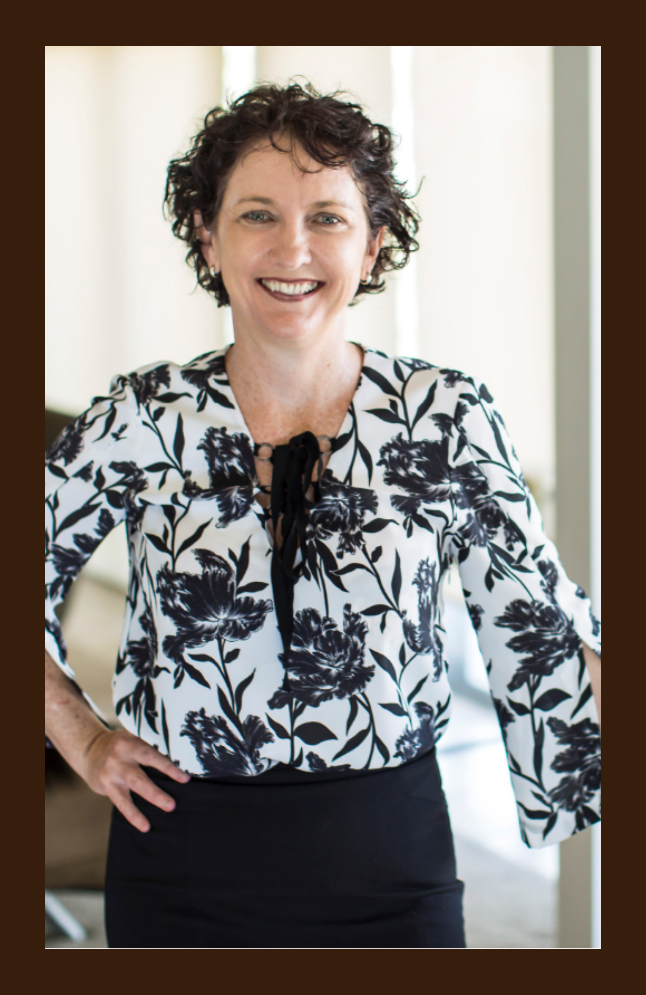 A woman with short curly dark hair is smiling and standing with one hand on her hip. She is wearing a white blouse with black floral print and a black ribbon tie, paired with a black skirt. The background is softly lit and appears to be an indoor setting.