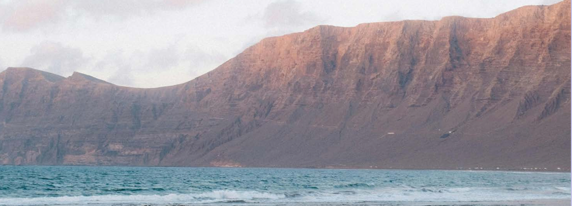 A view of a body of water with small waves, and a mountain range in the background under a cloudy sky.