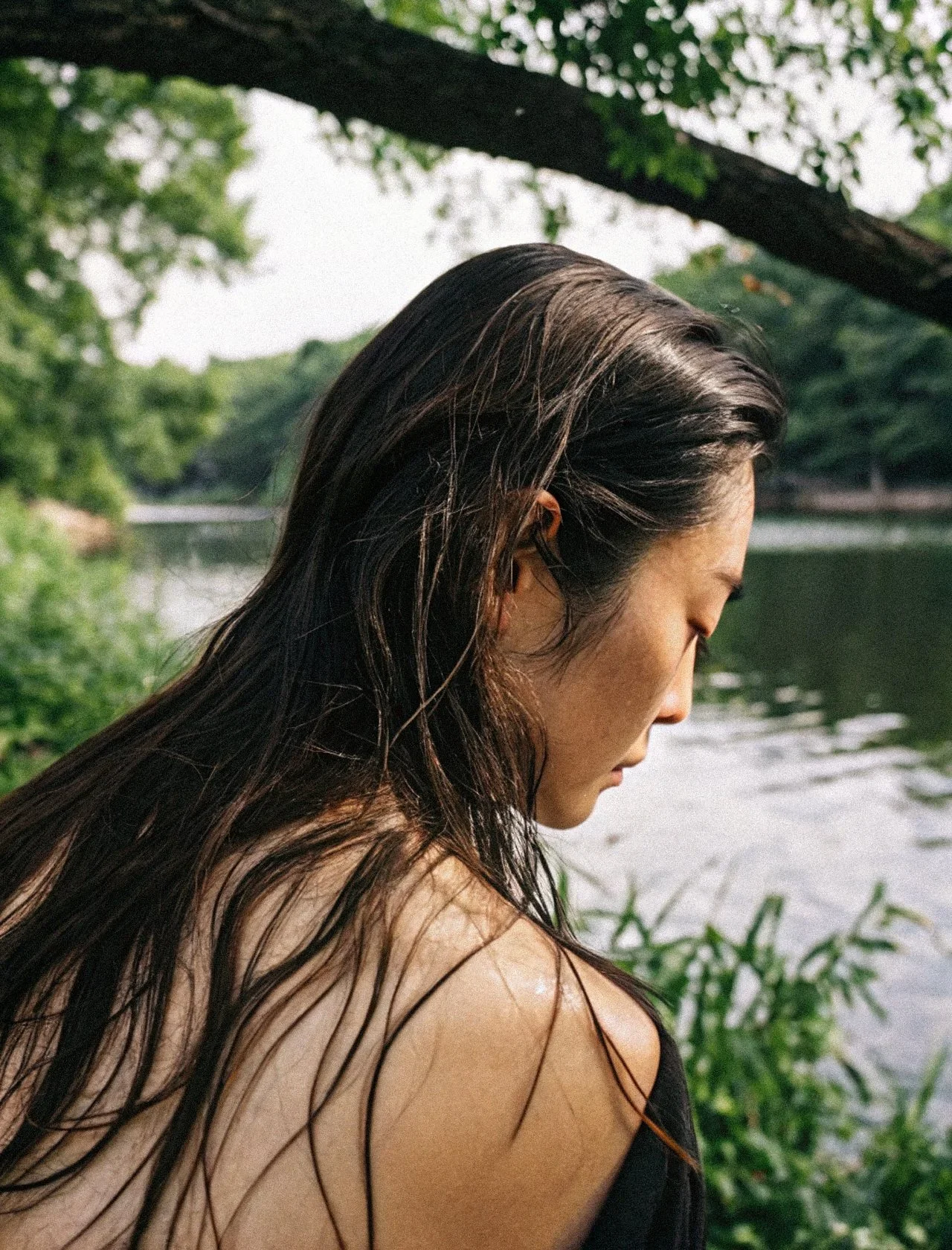 A woman with long, dark, wet hair sitting outdoors near a body of water, with trees and greenery around.
