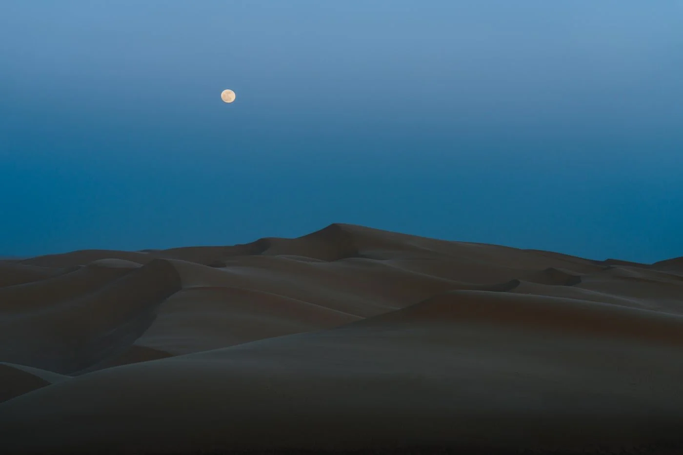 Dunes in a desert landscape at dusk with a visible full moon in the sky.