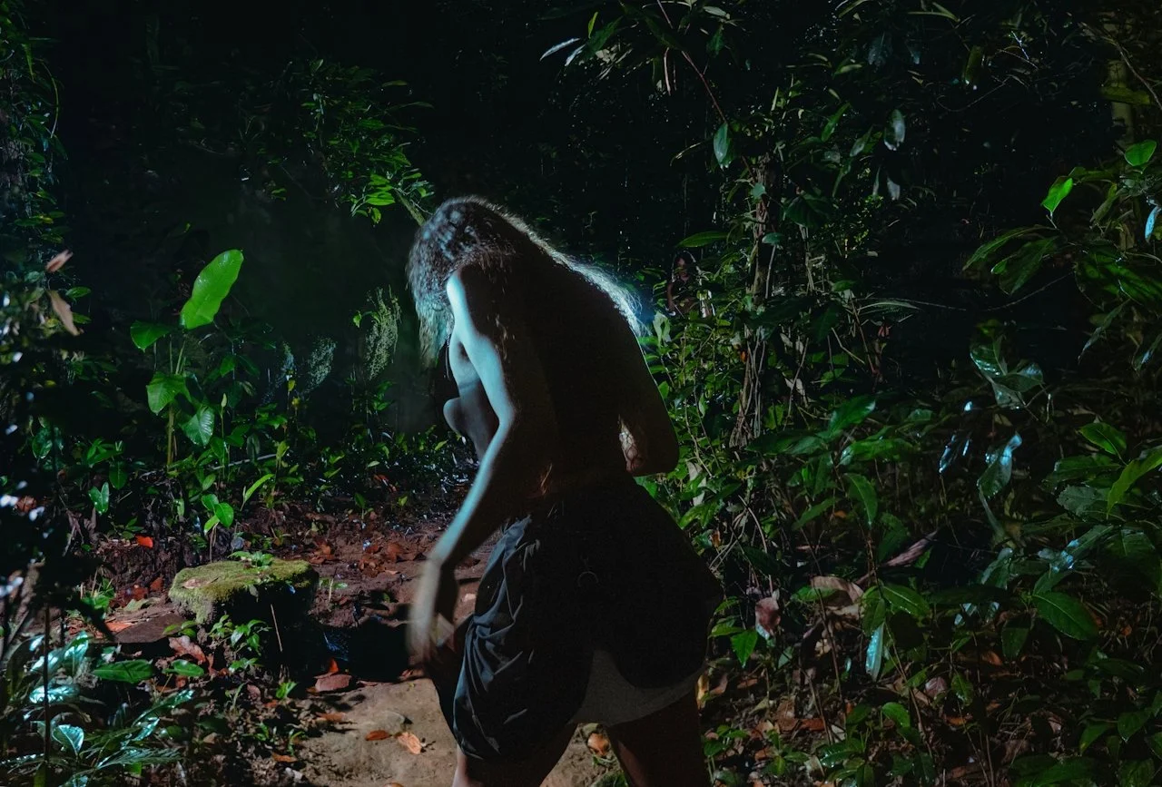 A woman crouching in a dense jungle at night, illuminated by a light behind her that highlights her silhouette and the surrounding foliage.