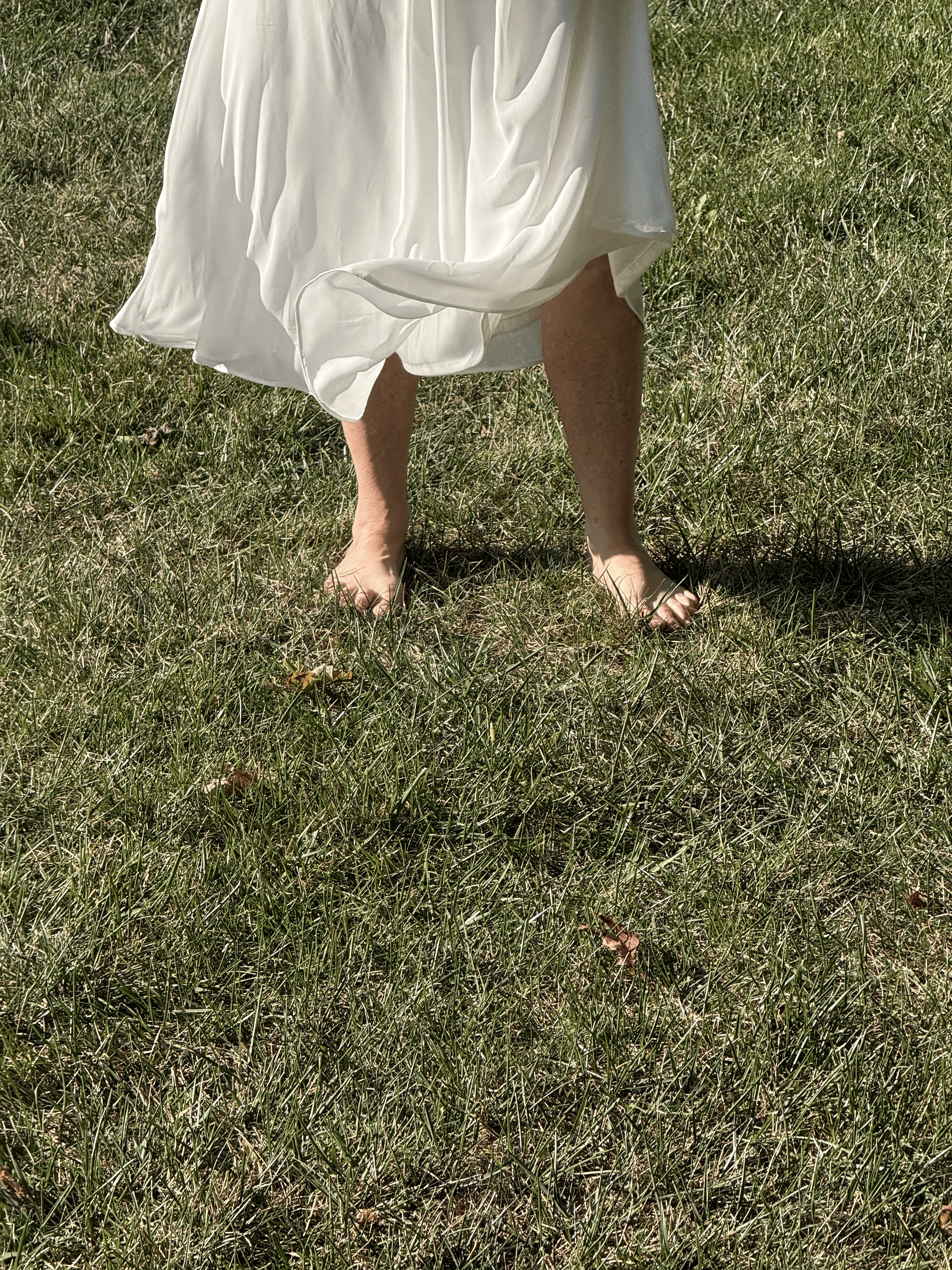 A person wearing a white dress standing barefoot on grass, with only their legs visible in the image.