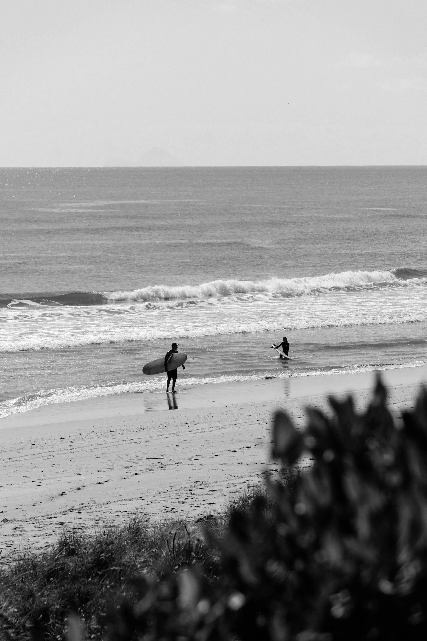 Two surfers on the beach with surfboards, one standing and walking towards the water, the other in the water with a surfboard, with ocean waves and a distant island in the background.