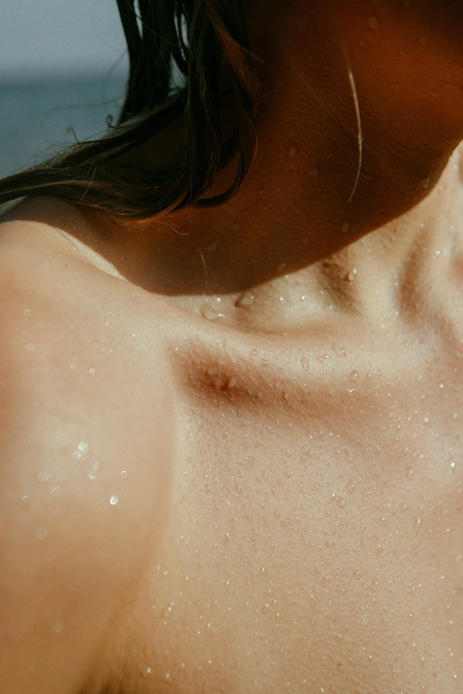 Close-up image of a person's shoulder, neck, and part of the face, with water droplets on skin and dark hair.