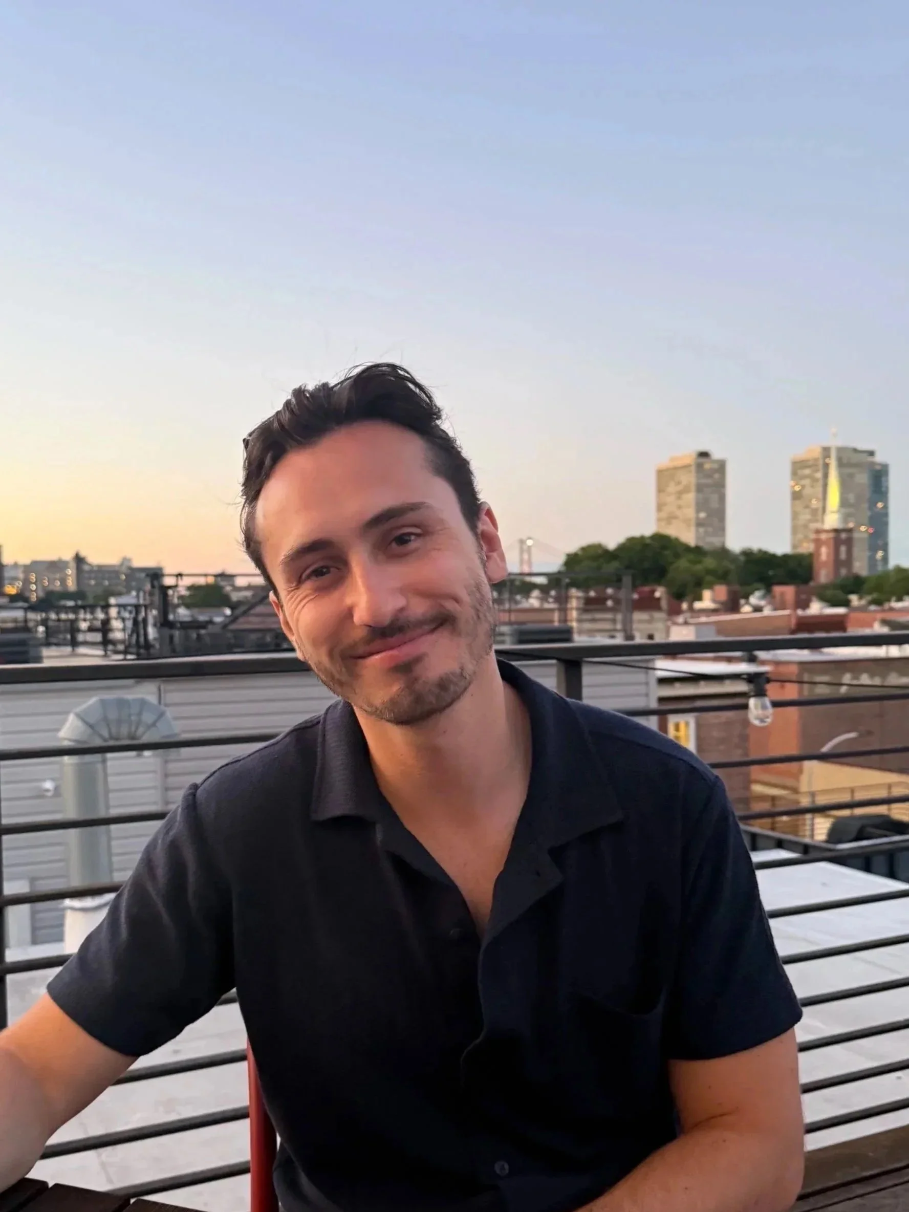 A smiling man with short dark hair and a beard sitting outdoors on a rooftop deck during sunset, with a cityscape of high-rise buildings and trees in the background.