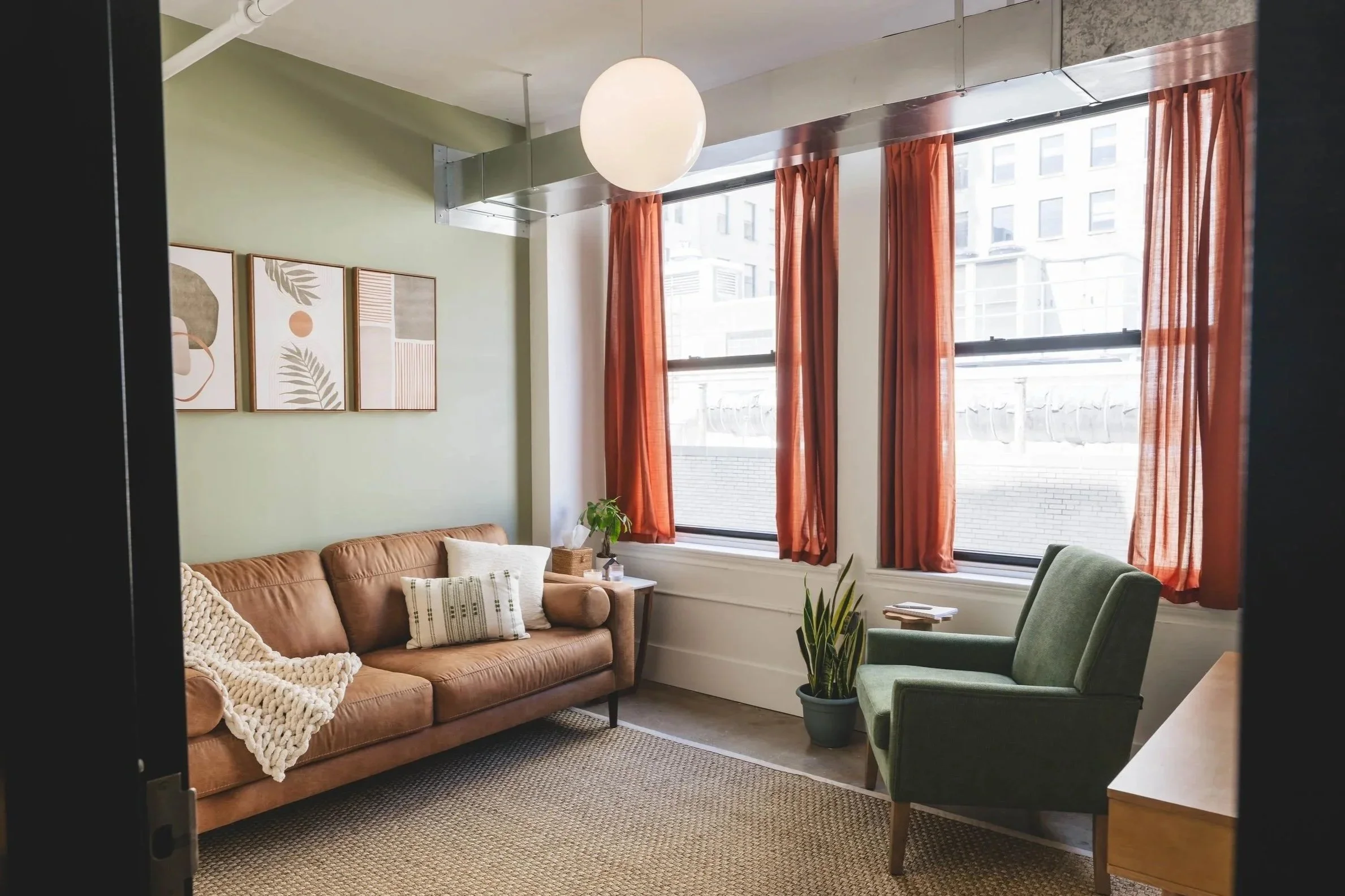 A cozy living room with a tan sofa, a white chunky blanket, and decorative pillows. There is a banana leaf plant in a gray pot, a small side table, and a green armchair. The room has large windows with orange curtains, and three framed art pieces hang on a light green wall. A round hanging light fixture is above, and part of a kitchen counter is visible on the right.