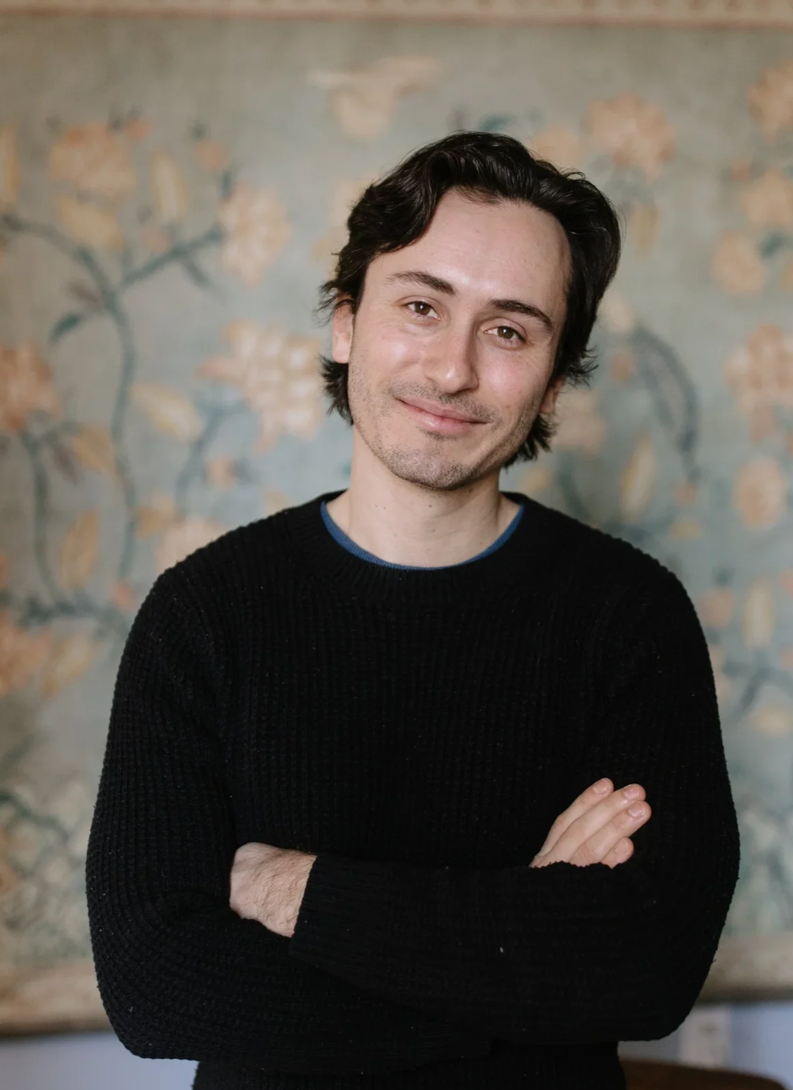 A young man with dark, wavy hair and light facial hair, wearing a black sweater over a blue shirt, smiling with arms crossed, standing in front of a floral-patterned background.