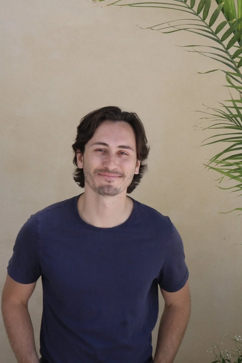 A man with dark hair and a goatee smiling, wearing a navy blue t-shirt, standing indoors near a beige wall and green potted plant.