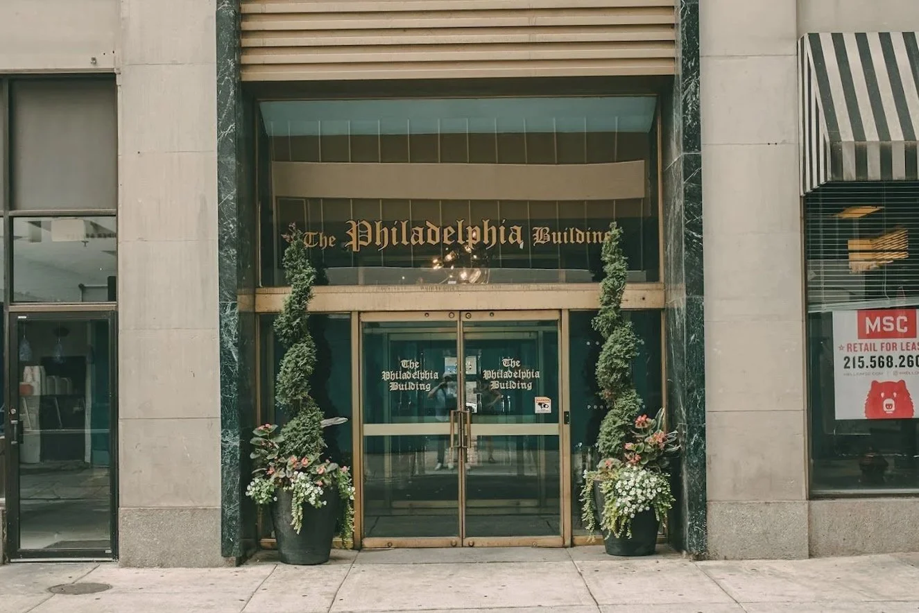 Entrance to The Philadelphia Building with glass doors, flanked by potted plants, and signage displaying the building's name in gold lettering.