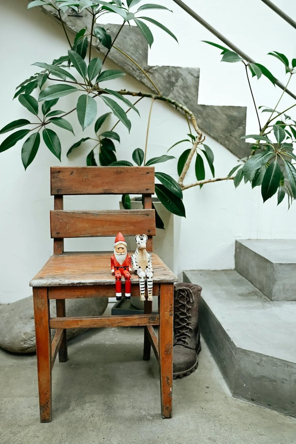 A wooden chair with a small Santa Claus and a zebra doll sitting on it, positioned near a large green plant and concrete stairs.