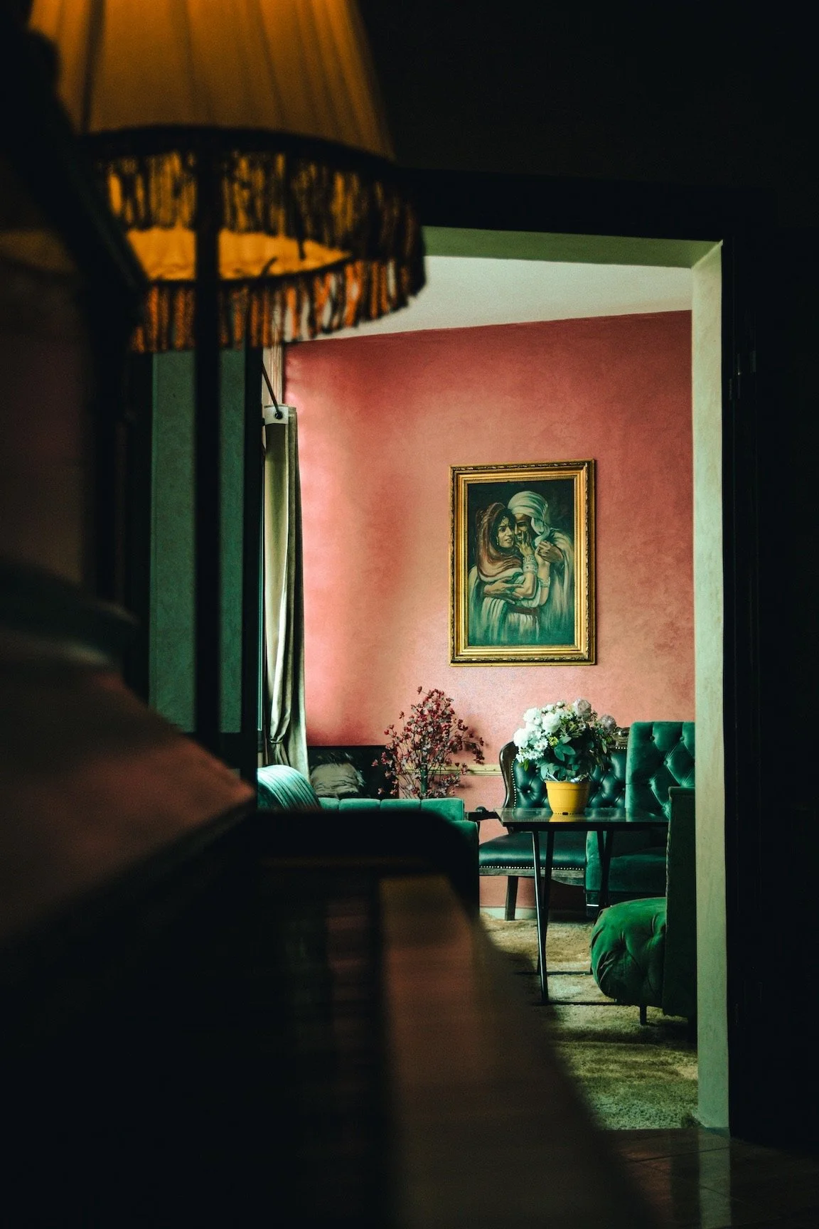 A cozy living room with pink walls, green furniture, and a framed portrait of a woman with a turban. There are flowers on the table and a lamp with a fringed shade in the foreground.