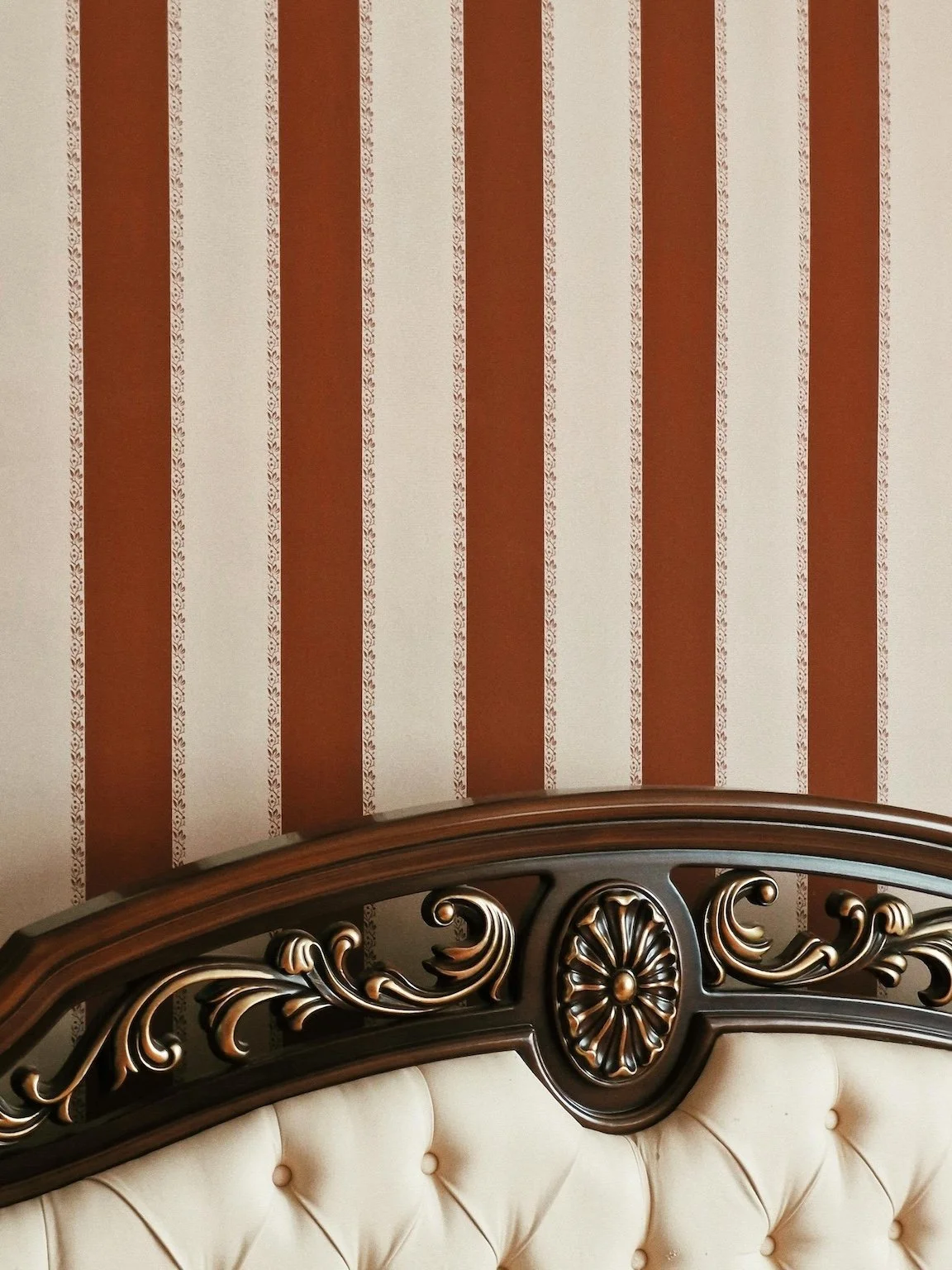 Part of a decorative headboard with carved wood design and cream-colored tufted upholstery, visible against a wallpapered wall with vertical stripes and floral patterns.