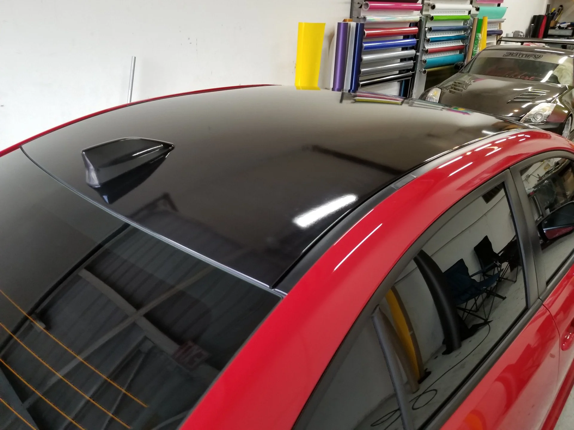 Close-up of a red car with a black roof and a shark fin antenna, parked indoors alongside another silver car, with shelves of colorful rolls of vinyl film in the background.