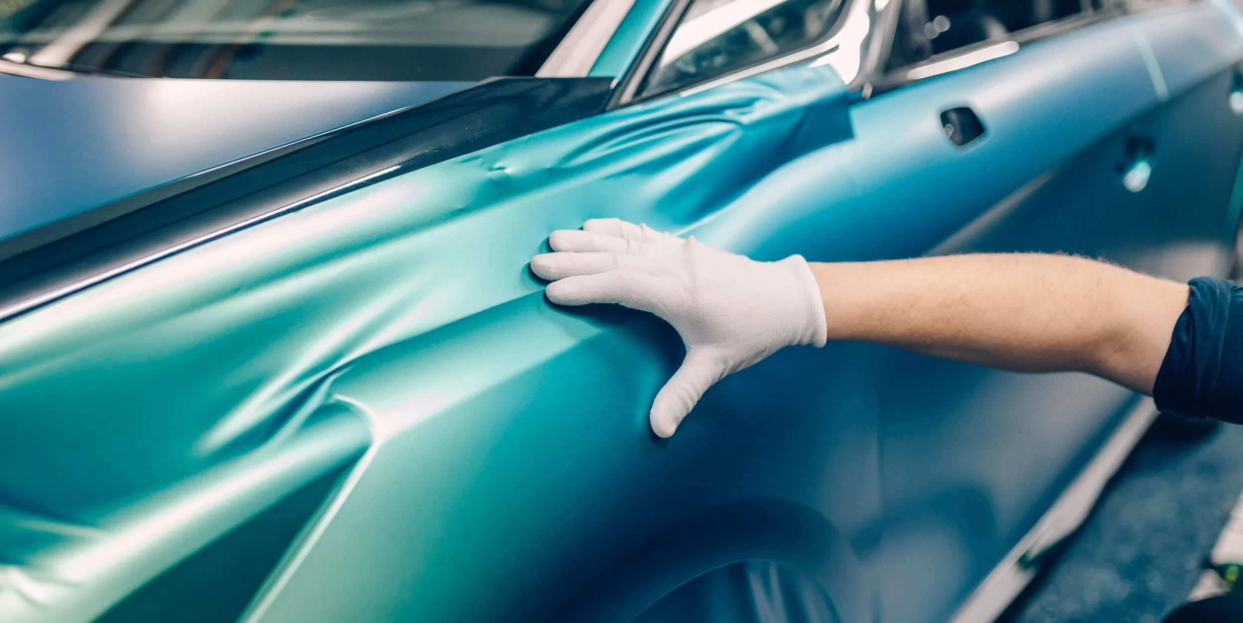 Person wearing white gloves applying a teal-colored vinyl wrap to a car's side panel.