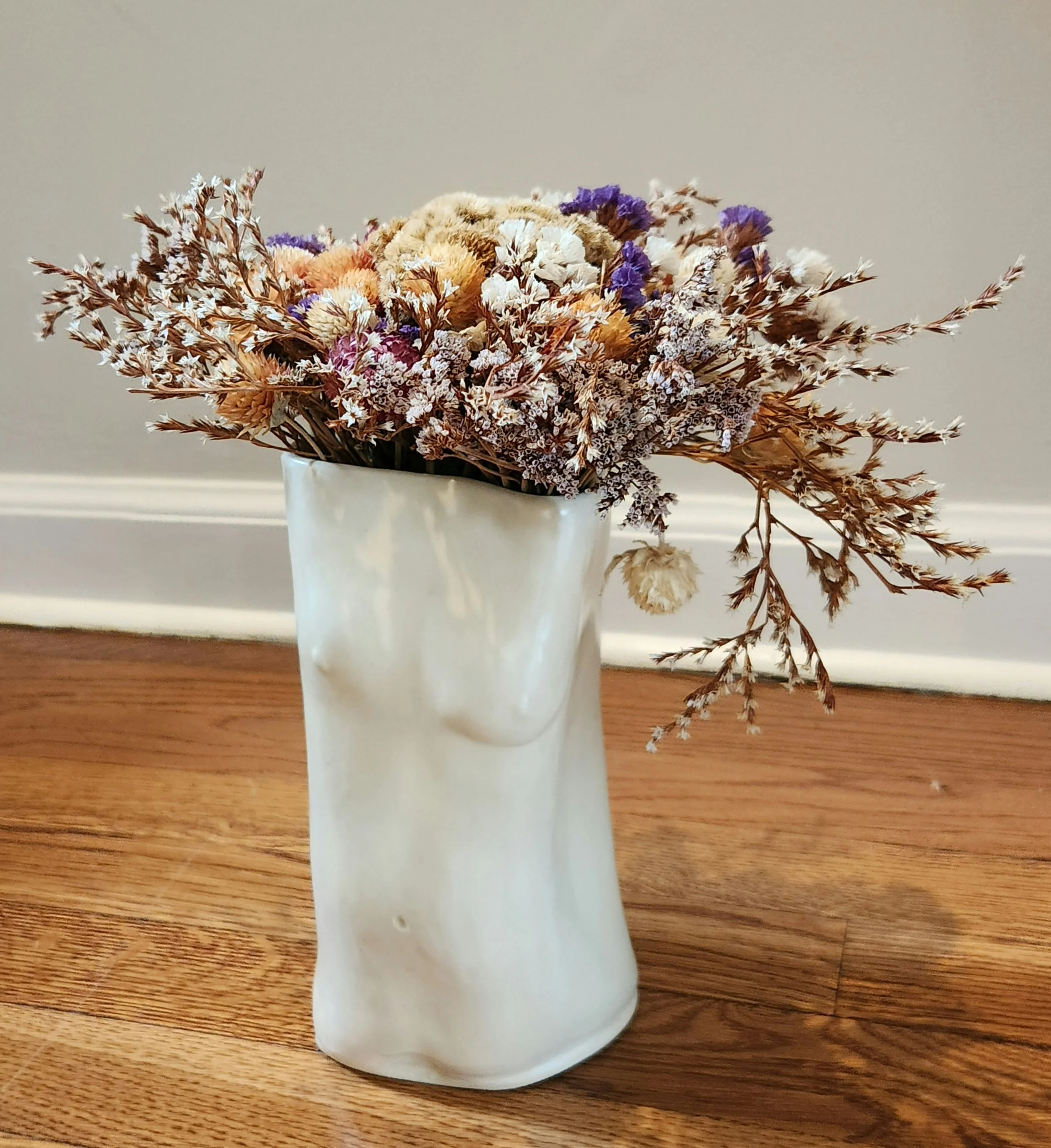 A ceramic vase shaped like a human face with closed eyes, containing a bouquet of dried flowers on a wooden floor.