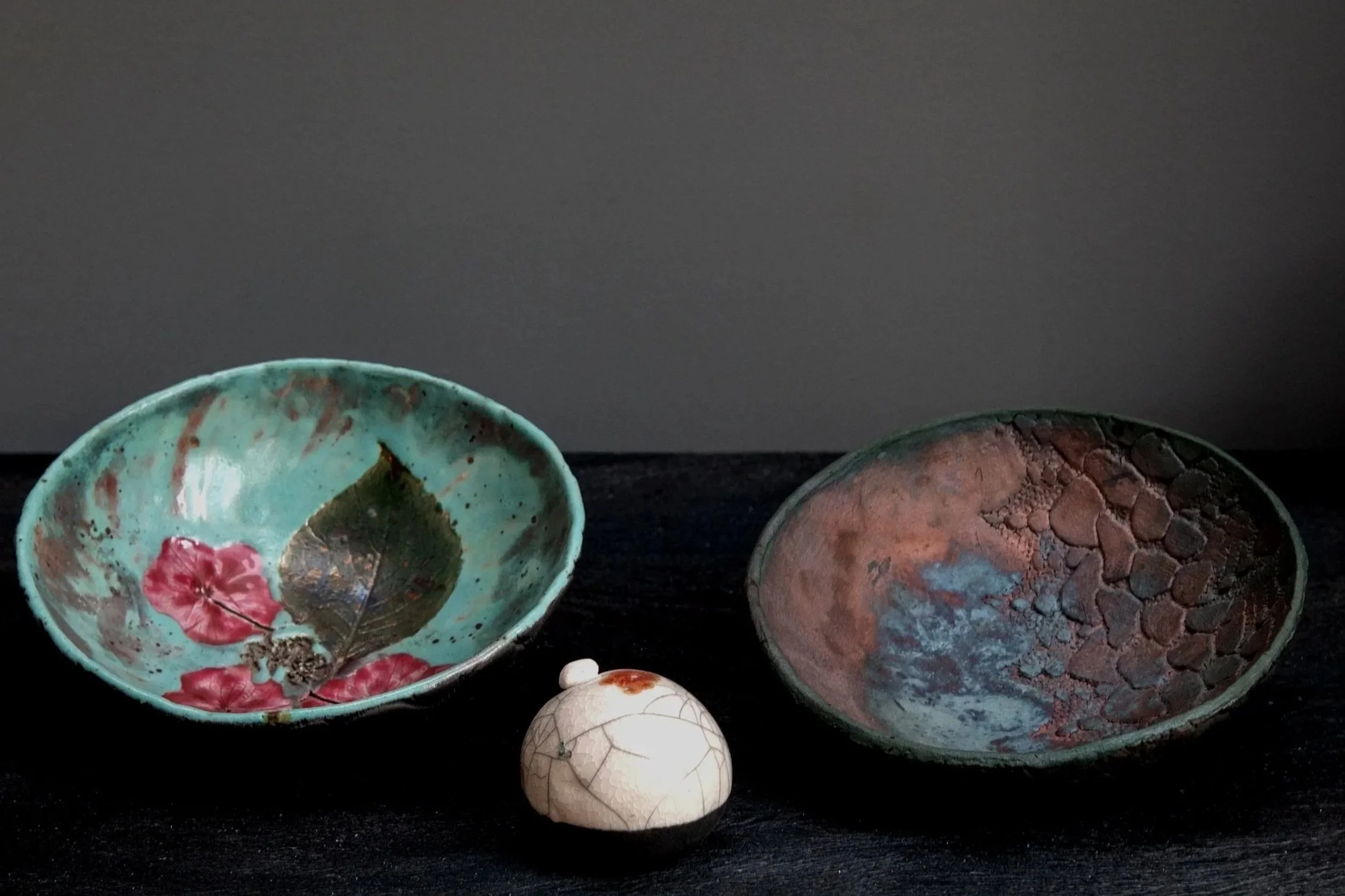 Decorative ceramic bowls on a dark surface, with one bowl containing a leaf and pink flower, and a small round ceramic object in front of them.