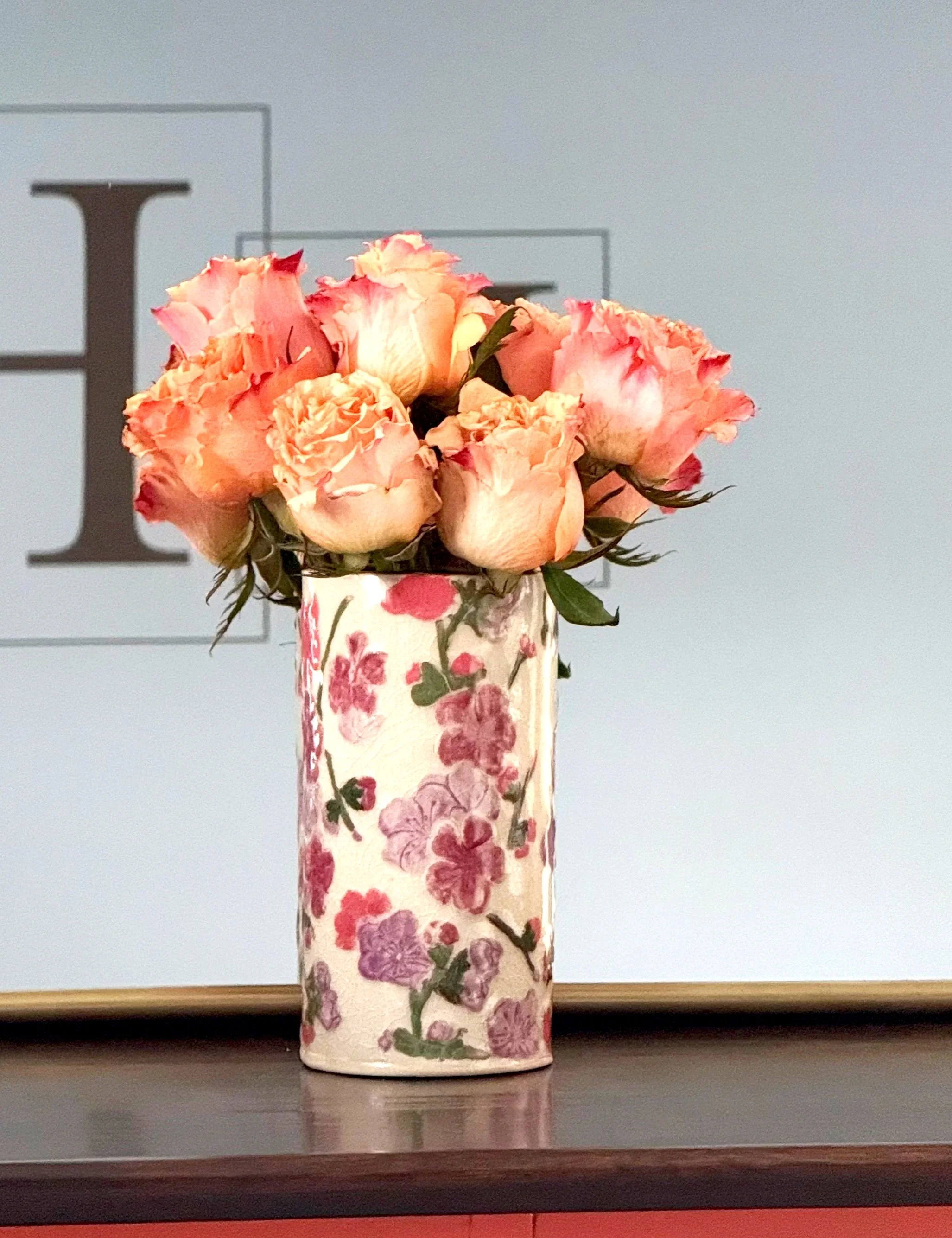 A bouquet of pink and peach roses in a white vase with pink and purple flowers pattern, placed on a dark surface.