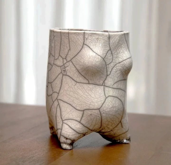 A ceramic cup with a cracked, marbled glaze pattern sitting on a wooden surface with sheer curtains in the background.