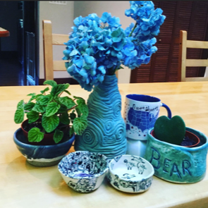 Arrangement of decorative items on a table including blue hydrangea flowers in a textured blue vase, a potted mint plant, a ceramic mug with a blue pattern, a green heart-shaped object, a green ceramic dish with the word 'BEAR,' and two small patterned bowls.