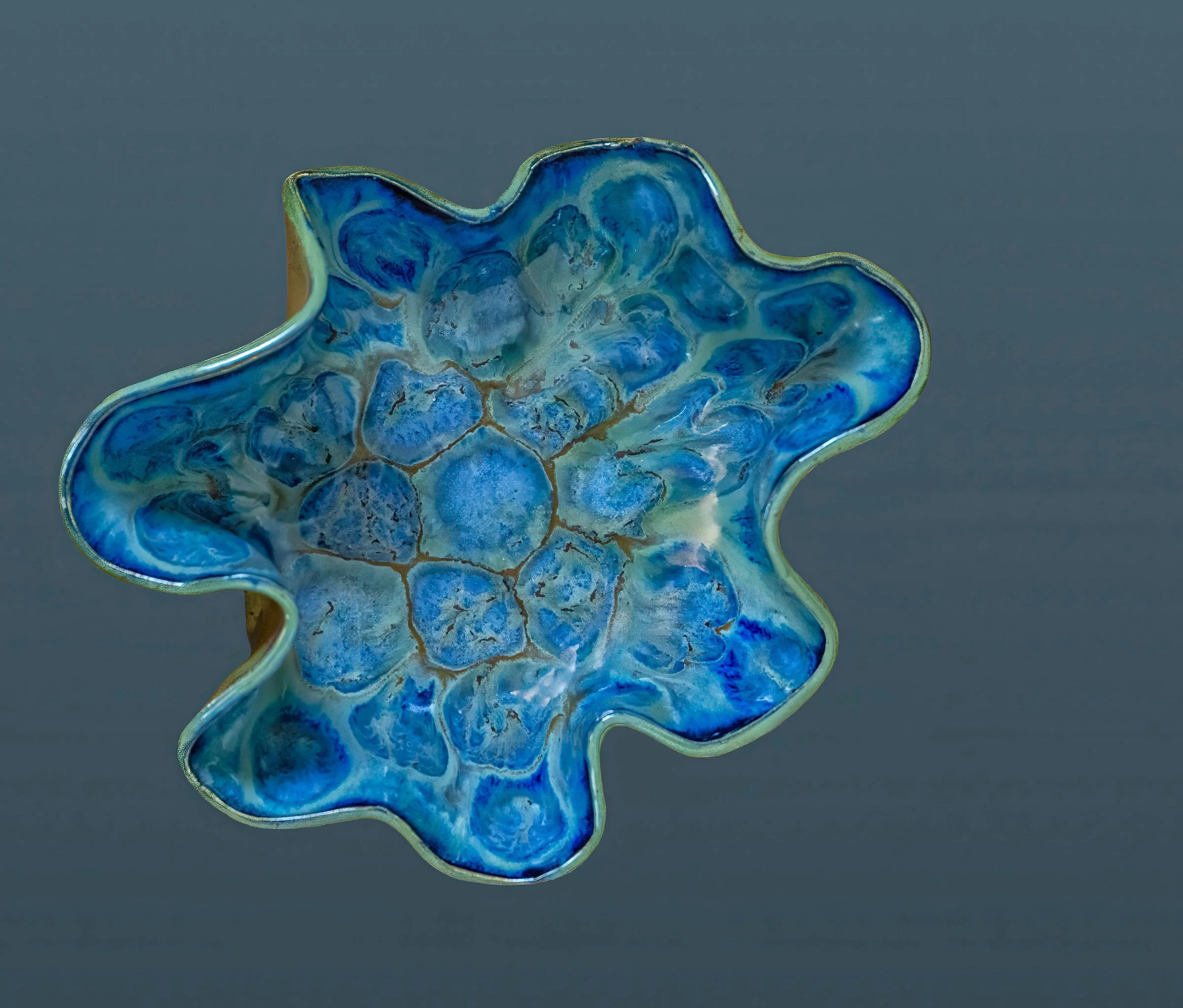 Close-up of a decorative ceramic bowl with blue and green marbled glaze, irregularly shaped with a ruffled edge against a plain grey background.