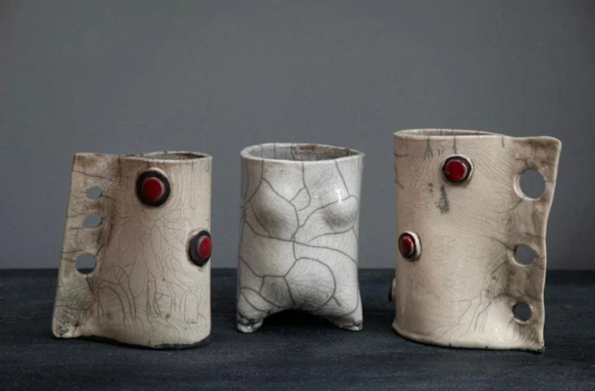 Three ceramic cups with an irregular cracked glaze finish, featuring round red embellishments and holes on the side, set against a plain gray background.
