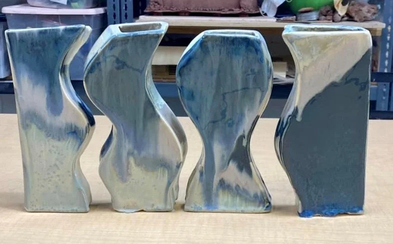 Four abstract ceramic vases with curved, wavy shapes and blue-gray marbled glaze, placed on a wooden surface in a workshop setting.