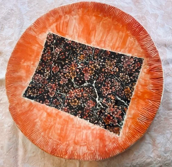 Decorative round plate with an orange rim and a black rectangular section in the center featuring a floral pattern with pink and orange flowers.