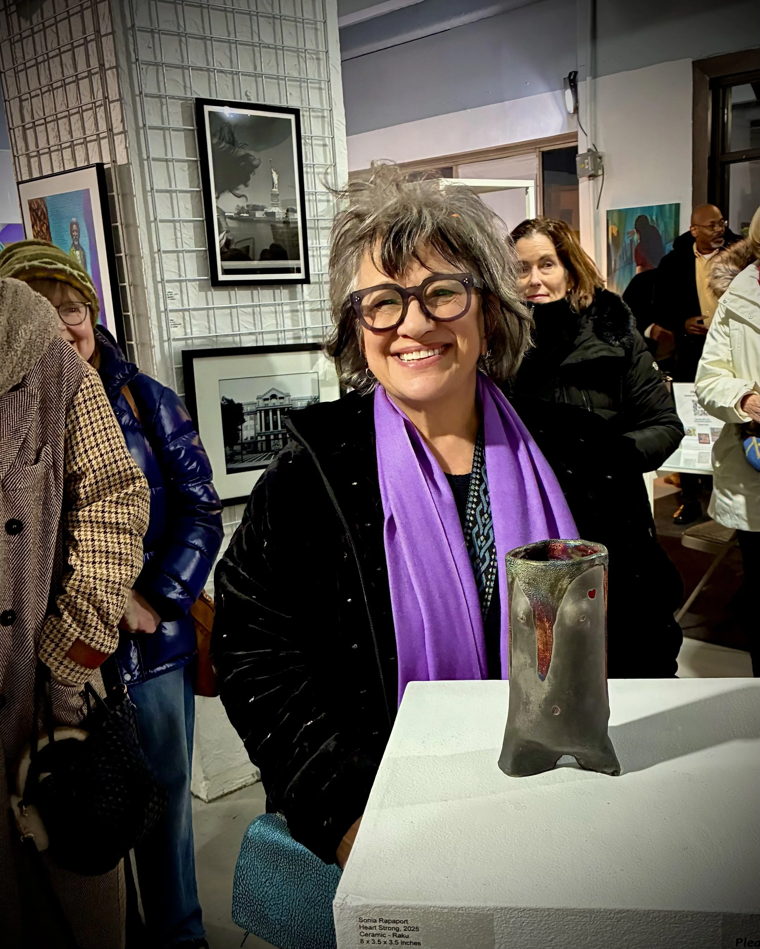 A woman with gray hair, glasses, and a purple scarf is smiling at an art gallery, standing next to a ceramic sculpture on a pedestal.