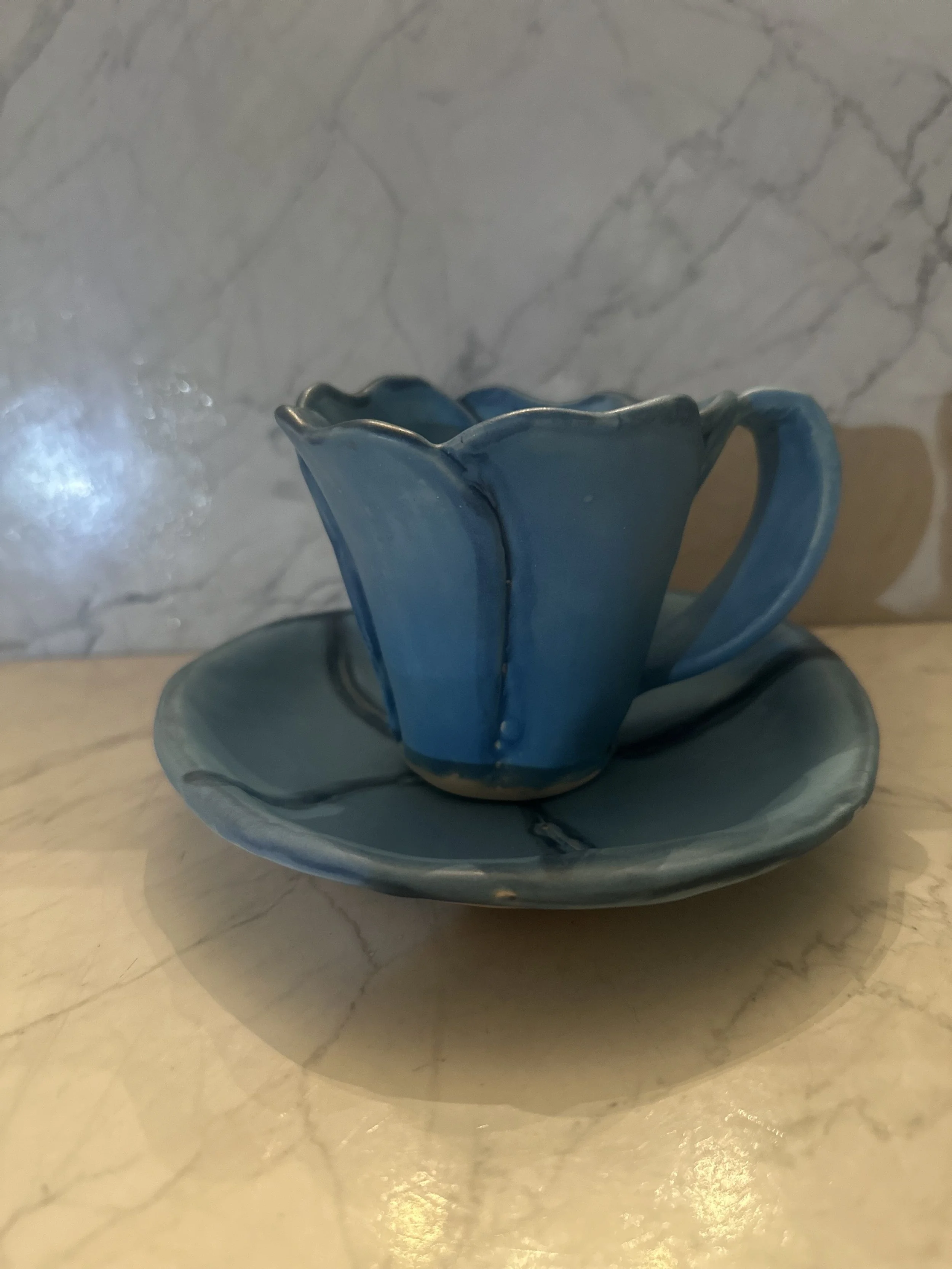 A blue ceramic teacup with a matching saucer, set on a marble surface with a marble wall backdrop.