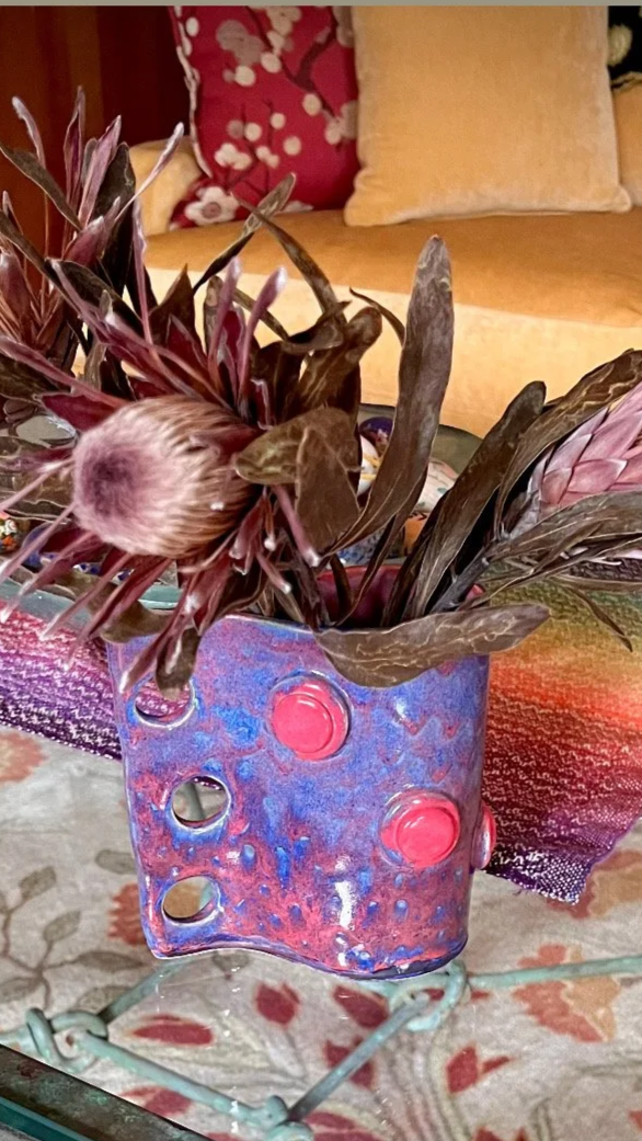 Decorative dried flower arrangement in a colorful, hole-pocked glass vase on a glass table, with a floral-patterned rug underneath and a beige couch with a patterned pillow in the background.