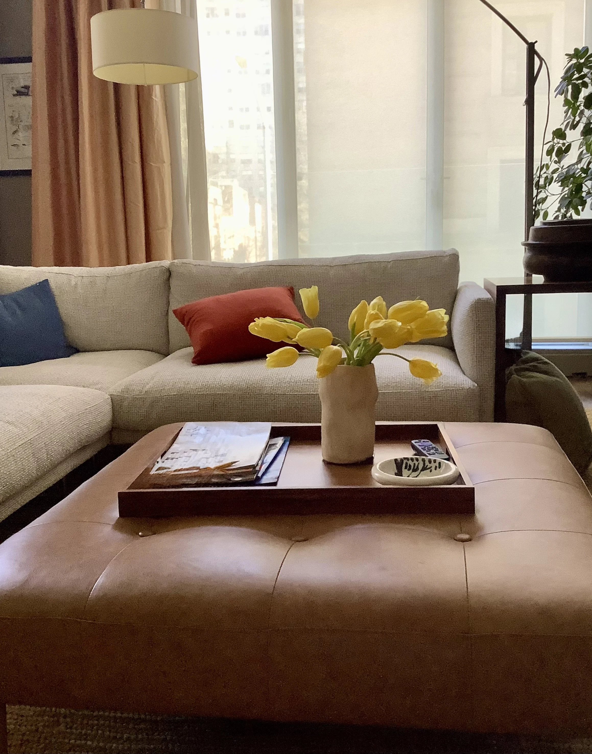 Living room with beige sofa, red pillow, yellow tulips in a vase on a wooden coffee table, magazines, and a remote, with large windows and a floor lamp.