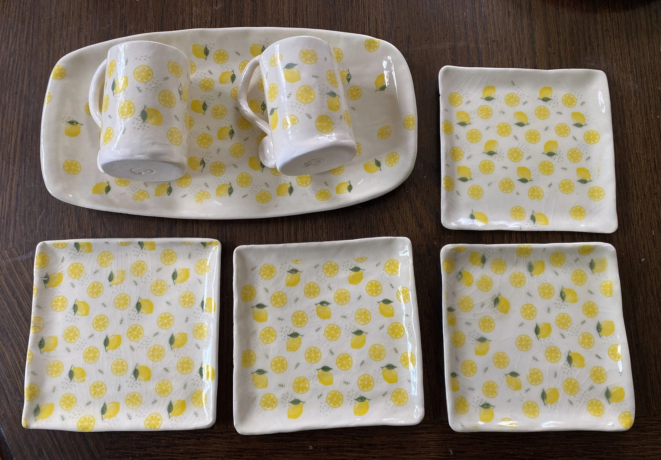 A set of for lemon-themed ceramic tableware with matching yellow lemon pattern on a dark wooden table. Includes a rectangular tray with two matching cups, two square plates, and two smaller square plates.