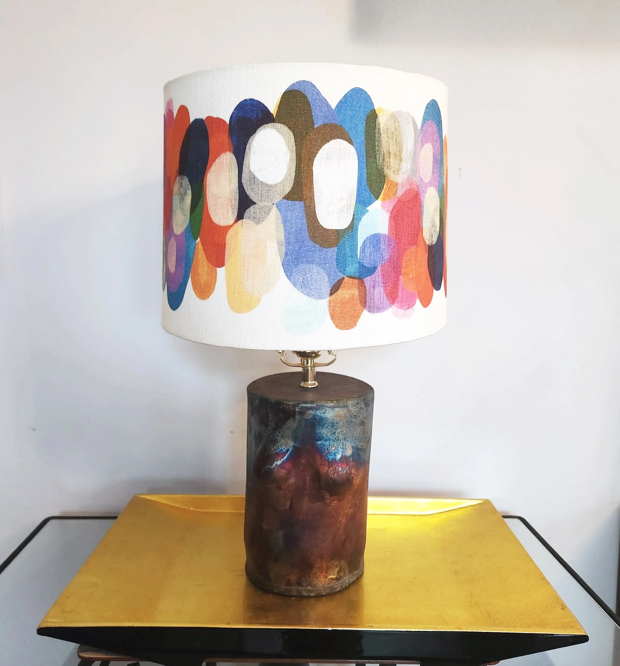 A table lamp with an abstract, colorful oval pattern lampshade and a textured, multicolored ceramic base, placed on a yellow surface.