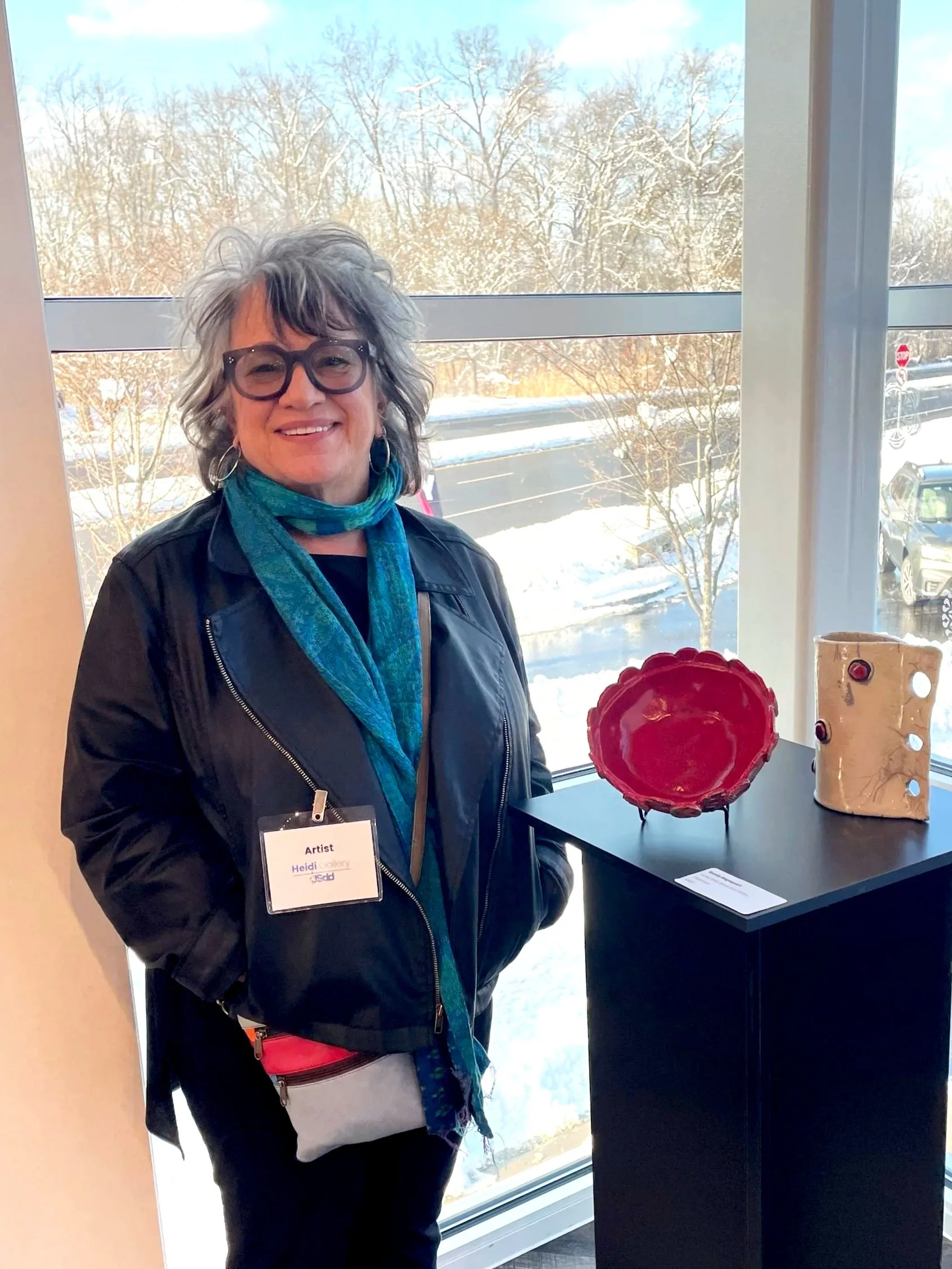 A woman with gray hair, glasses, and a black jacket standing indoors beside a display table with ceramic art pieces. Outside the window is a snowy landscape with trees and parked cars.
