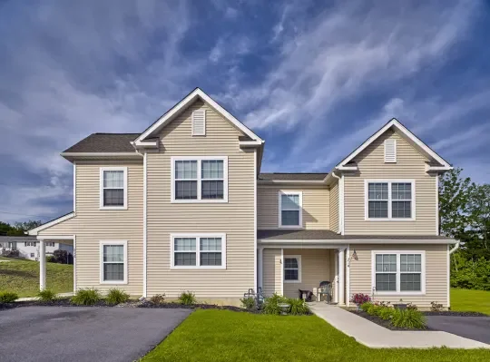 A two-story beige house with multiple windows and a small front porch, with a lawn and paved driveway under a partly cloudy sky.